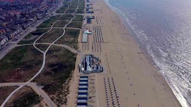 Wide horizontal aerial drone video of Katwijk aan Zee beachfront resort, North Sea foam waves, sun glint reflections, sand dunes, and town skyline in South Holland, Netherlands on sunny afternoon