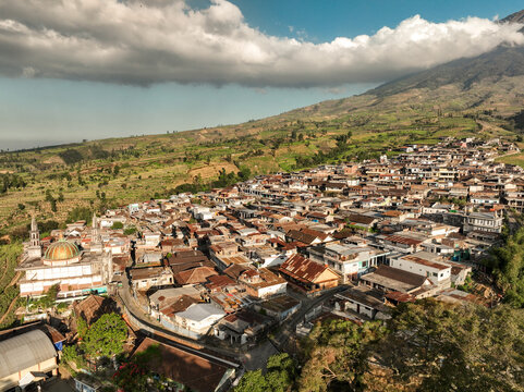 Aerial view of Kruwisan village with its golden-domed mosque, dense rooftops, and terraced agricultural fields set against a mountainous landscape in Kledung, Central Java, Indonesia.