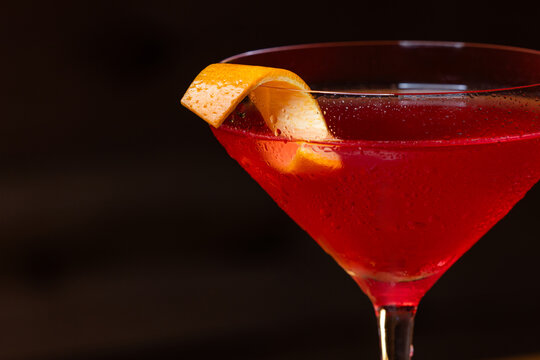Pink cosmopolitan cocktail in coupe glass. An orange twist adorns the rim. Dark wood table and dark background create a rustic bar atmosphere.