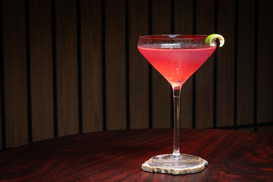 Pink cosmopolitan cocktail in martini coupe glass. Lime twist adorns the rim. Dark wood table create depth. Sleek, elegant, sophisticated mood