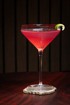 Pink cosmopolitan cocktail in martini coupe glass. Lime twist adorns the rim. Dark wood table create depth. Sleek, elegant, sophisticated mood