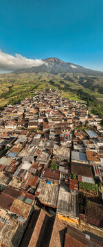 Aerial view of Kruwisan village nestled among lush green hills and mountains with dense rooftops under a clear blue sky in Kledung, Central Java, Indonesia.