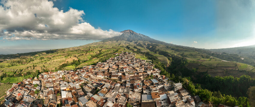 Aerial view of Kruwisan village at the foot of Mount Sindoro, surrounded by lush terraced agricultural fields and rolling hills under a blue sky in Kledung, Central Java, Indonesia.