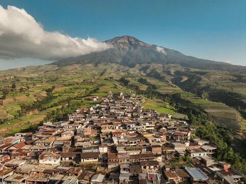 Aerial view of Kruwisan village nestled at the base of Mount Sumbing with dense residential rooftops and lush terraced agricultural fields in Kledung, Central Java, Indonesia.