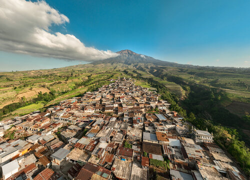 Aerial view of Kruwisan village nestled in the lush agricultural landscape at the base of a majestic volcano under a clear blue sky in Kledung, Central Java, Indonesia.