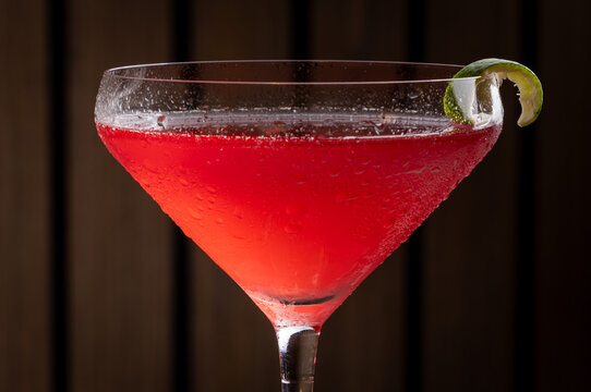 Pink cosmopolitan cocktail in martini coupe glass. Lime twist adorns the rim. Dark wood table create depth. Sleek, elegant, sophisticated mood