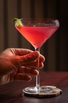 Pink cosmopolitan cocktail in martini coupe glass. Lime twist adorns the rim. Dark wood table create depth. Sleek, elegant, sophisticated mood