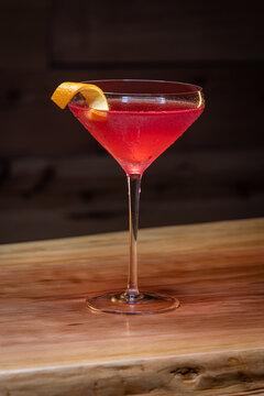 Pink cosmopolitan cocktail in coupe glass. An orange twist adorns the rim. Dark wood table and dark background create a rustic bar atmosphere.