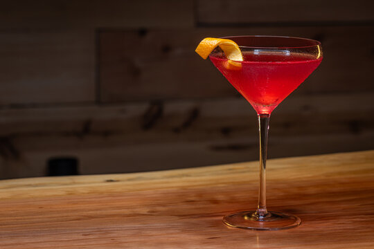 Pink cosmopolitan cocktail in coupe glass. An orange twist adorns the rim. Dark wood table and dark background create a rustic bar atmosphere.