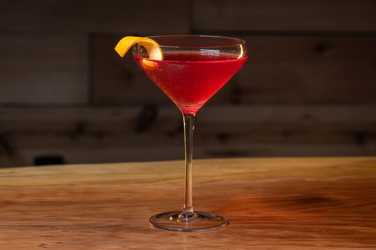 Pink cosmopolitan cocktail in coupe glass. An orange twist adorns the rim. Dark wood table and dark background create a rustic bar atmosphere.