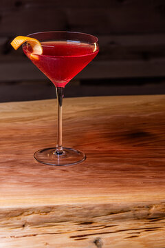 Pink cosmopolitan cocktail in coupe glass. An orange twist adorns the rim. Dark wood table and dark background create a rustic bar atmosphere.