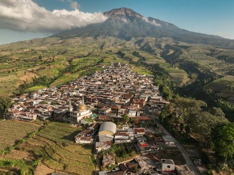 Aerial view of Kruwisan village nestled at the base of Mount Sindoro, featuring a mosque with a golden dome surrounded by terraced agricultural fields in Kledung, Central Java, Indonesia.