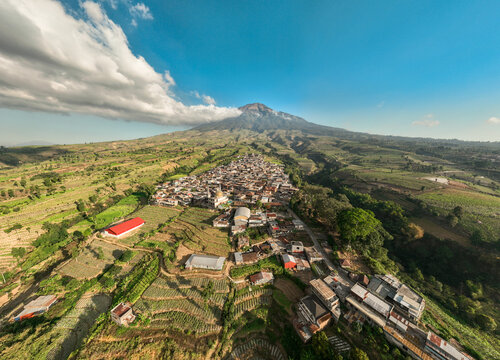 Aerial view of Kruwisan village nestled among terraced agricultural fields with a majestic volcano in the background under a clear blue sky in Kledung, Central Java, Indonesia.