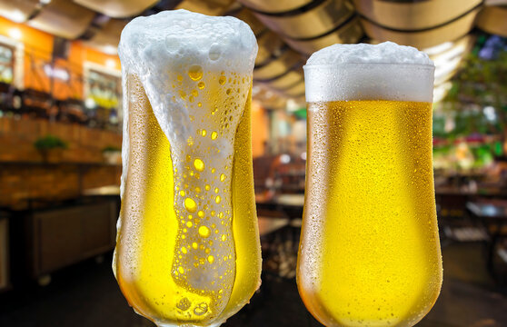 Two frosty glasses of cold beer with thick overflowing foam, set against the warm, blurred background of a pub.