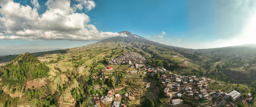 Aerial view of Kruwisan village nestled among lush terraced fields and rolling hills at the base of a majestic mountain under a clear blue sky in Kledung, Central Java, Indonesia.