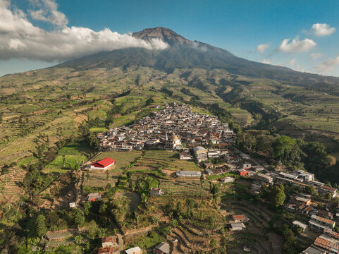 Aerial view of Kruwisan village nestled at the foot of a majestic mountain surrounded by lush green terraced fields and agricultural land in Kledung, Central Java, Indonesia.