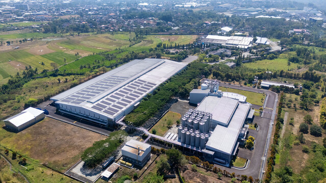 Aerial view of a large industrial factory complex featuring solar panels on the roof, tall storage silos, and surrounding green fields and trees in Salatiga, Central Java, Indonesia.