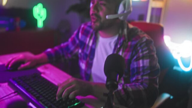 Man wearing headset grips mouse and clenches fist at keyboard desk with neon signs in studio; competitive concentration.
