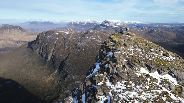 Aerial view of Ben Mor Coigach featuring rugged snow-dusted rocky peaks and a lone hiker standing on the summit under a clear blue sky in Ullapool, Scotland, United Kingdom.