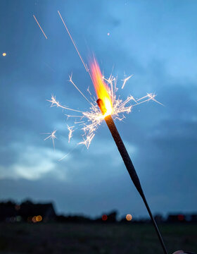 Sparking firework stick glowing against blue evening sky with bright sparks and blurred city lights
