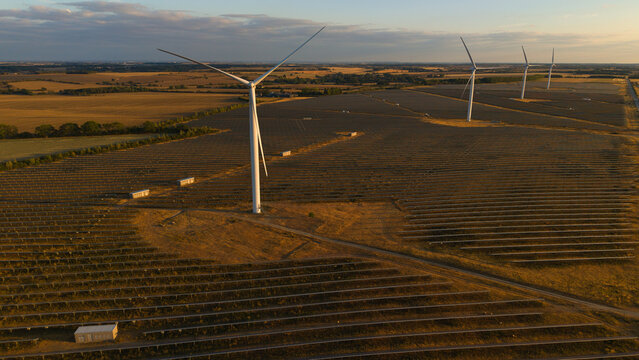 Aerial view of wind and solar renewable energy park at sunset with rows of solar panels and wind turbines in the countryside of Chelveston, England, United Kingdom.