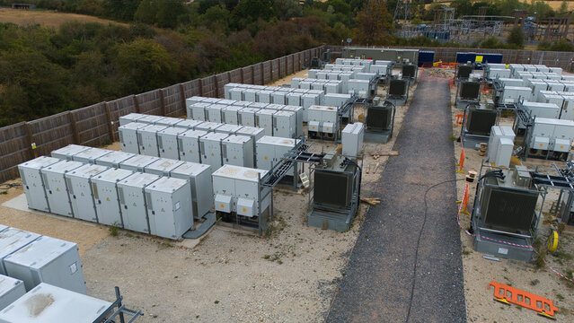 Aerial view of a Battery Energy Storage facility and Substation with rows of grey electrical cabinets, gravel pathways, and a wooden perimeter fence in Stony Stratford, England, United Kingdom.