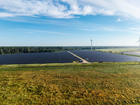 Aerial view of a large solar farm with rows of photovoltaic panels, surrounded by fields, with wind turbines in the distance under a blue sky in Targale, Ventspils Municipality, Latvia.