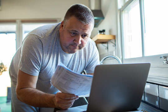Middle-aged man checking household bills on laptop in home kitchen