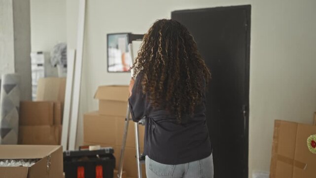 Woman smiling while holding boxes and a paint roller in a studio among moving supplies and stacked cardboard; home settling contentment.