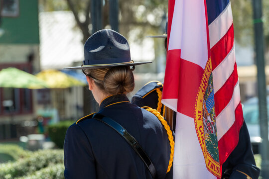 A female police officer stands erect at a parade wearing a campaign hat, a navy blue dress uniform with an orange cord over her right shoulder. Her long hair is in a slicked-back bun. 