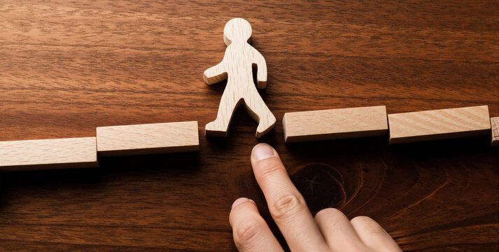 Wooden figure crosses gap on block bridge. Hand guides figure over obstacle. Problem solving, transition, progress. Represents overcoming challenges. Strategic pathway. Determination aids development.