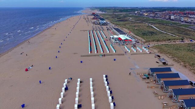 Wide horizontal aerial drone video of Katwijk aan Zee beach resort, white and red cabins, wooden holiday chalets North Sea waves sand dunes and town skyline in South Holland, Netherlands on afternoon