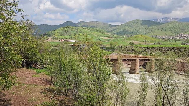 Old stone bridge arches over a swift, foaming river, framed by lush trees, with a distant town and snow-streaked mountains beneath dramatic clouds.
📍 Dorud, Sarvabad, Kurdistan 