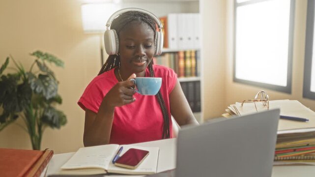 Woman holding coffee cup by hand while wearing headphones at desk with laptop, open books and glasses in a building; focused study.