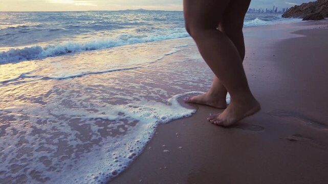 follow shot captures bare female legs walking along the shoreline into the water as foamy waves roll in during golden hour.