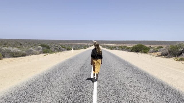 Woman with long brown hair walking away on a desert road under a clear blue sky in the middle of a dry rural outback region in Western Australia