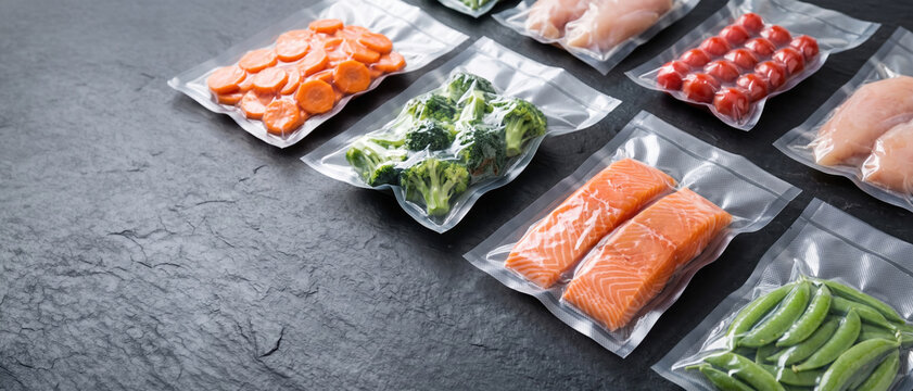 Various food items are packed in clear vacuum bags on a dark surface. The bags include chicken, salmon, vegetables, and fruits. This scene shows preparation for cooking or storage