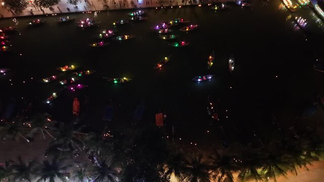 Illuminated boats drift on dark water at night in Hoi An, Vietnam, with palm trees and festive lights creating a vibrant scene