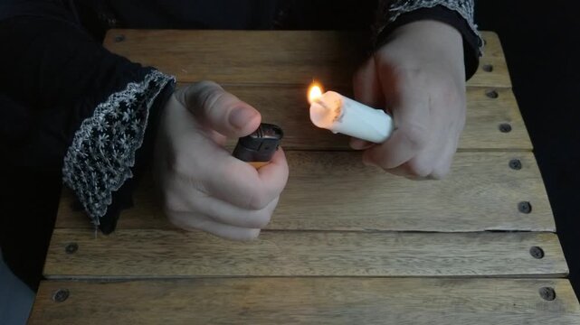 Close-up of a person's hand lighting a candle using a lighter. The scene shows a simple everyday activity involving an open flame and candle, suitable for themes related to lighting, household activit