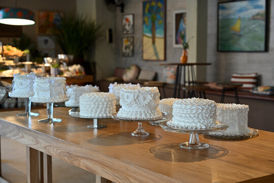 multiple white decorated cakes on glass stands on long wooden table, luxury brazilian event dessert buffet display with lounge backdrop, wide horizontal shot