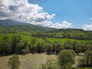 Obraz premium Wide muddy river flows past dense green trees, framed by vibrant fields, distant houses, and snow-streaked mountains beneath a partly cloudy sky. 📍 Dorud, Sarvabad, Kurdistan 