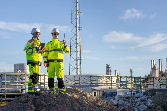 Engineers in safety gear checking project details on tablet in front of power plant architecture, Professional industrial team with digital tablet and walkie talkie outdoors at refinery plant