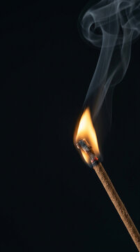 Close-up photograph of a burning matchstick with a bright orange flame, glowing ember, and delicate curling smoke against a deep black background. The dramatic composition provides strong negative spa