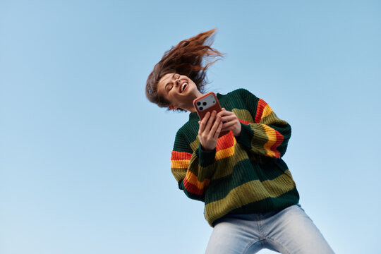 A joyful teen wearing a colorful striped sweater holds a smartphone outdoors, hair in motion, against a clear blue sky. Concept: connectivity, casual youth, carefree moments.