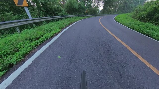 POV cycling through a winding mountain road surrounded by lush green forest, guardrails and a sharp curve, creating an immersive outdoor adventure scene.