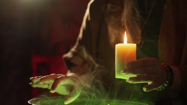A close-up of a woman's hands hovering over a cauldron. Green steam rises. A beeswax candle burns, and wax drips down. Preparing a potion
