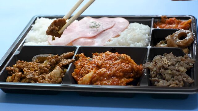Wooden chopsticks picking up fried meat from plastic lunch box against a blue background, side view.
