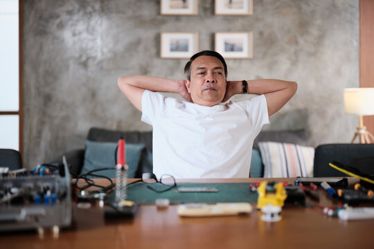 Mature Asian man concentrates on repairing home electrical appliance, disassembling and soldering circuit board, tired from precision detail focusing, taking a break, resting from his workshop hobby.