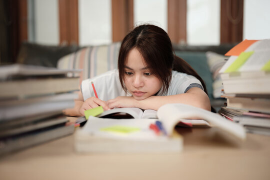 Asian teenage girl concentrates on pile of books, focuses on learning at home with pile of textbooks, reading and writing, self-study for school, Homeschooling, university education lifestyle.