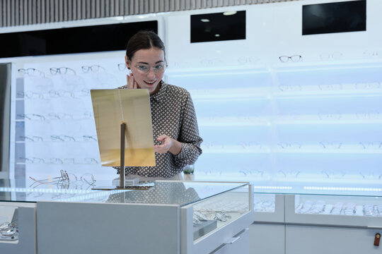 One attractive White female customer chooses and tries on eyeglasses, looks in mirror for improved eyesight for individual lifestyles, and makes decisions on options on store display in optical shop.